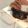 A black PU leather crossbody sling bag with gold hardware resting on a white canvas and a beige fabric.