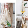 A hand holds a beige mesh grocery tote bag filled with produce, set against a light background.