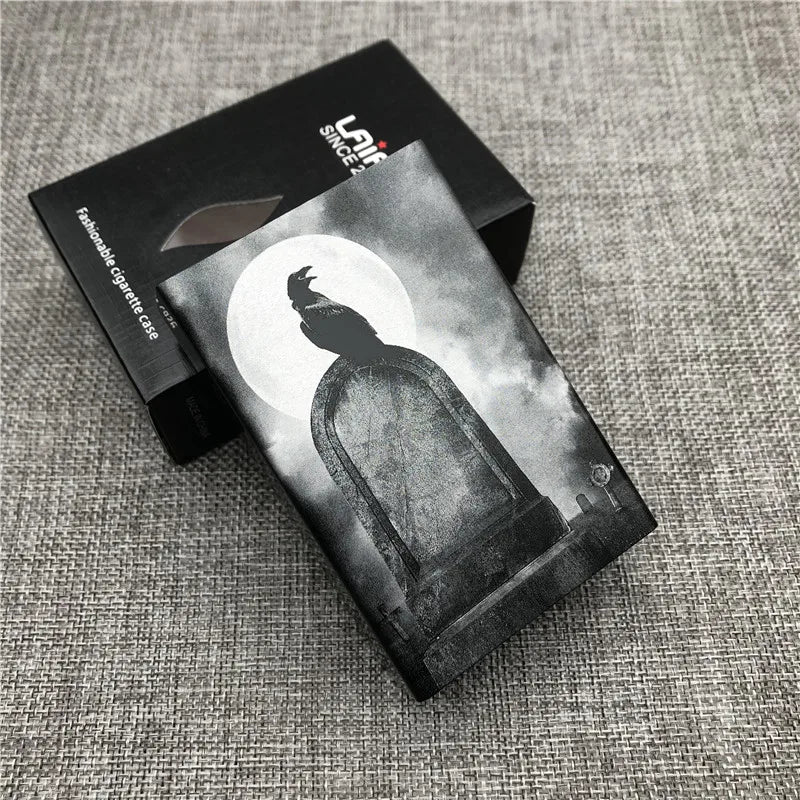 A black cigarette case with a silhouette of a bird against a moon background is displayed on a gray fabric surface.