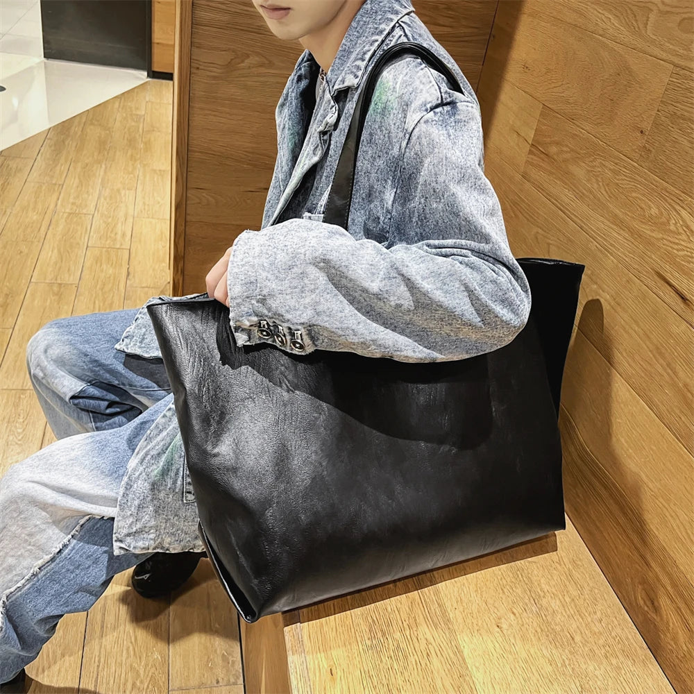 A person is sitting on the floor wearing jeans and a denim jacket, holding an oversized black PU leather tote bag.