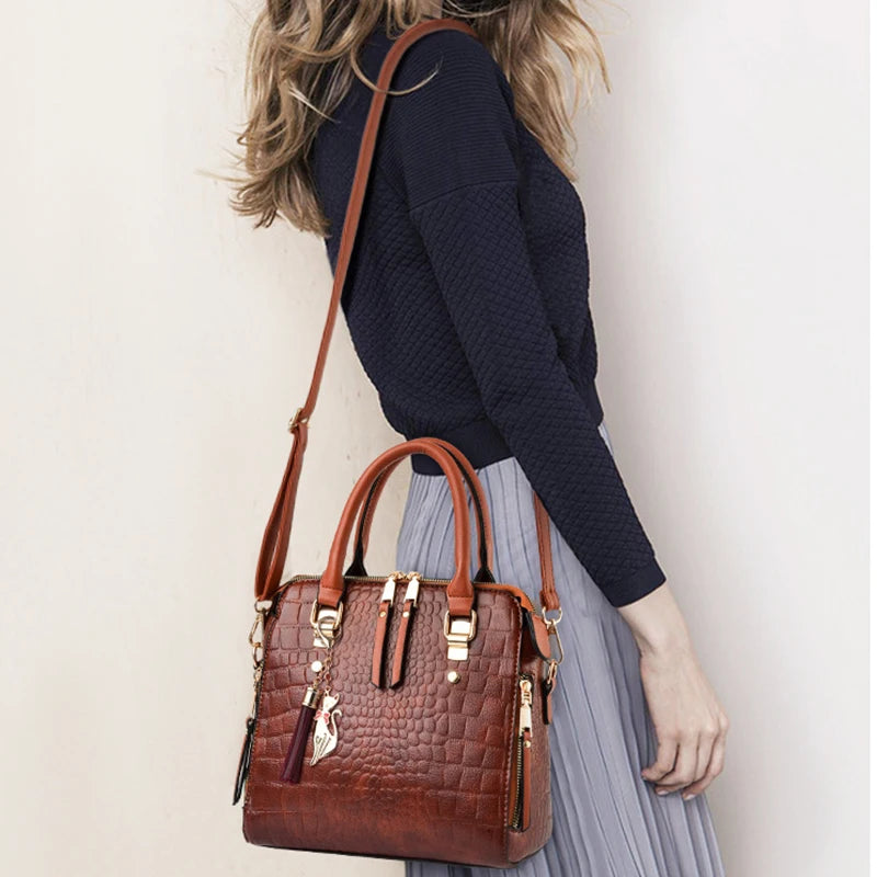 A brown crocodile-embossed leather tassel shoulder bag carried by a person wearing a dark blue sweater and light gray skirt.