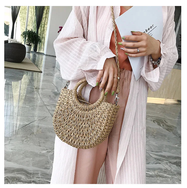 A woman holds a handwoven straw moon bag with a top handle while wearing a pink striped dress.