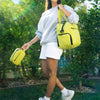 A woman wearing a light gray sweatshirt and white shorts is carrying a bright yellow large capacity travel duffle bag outdoor