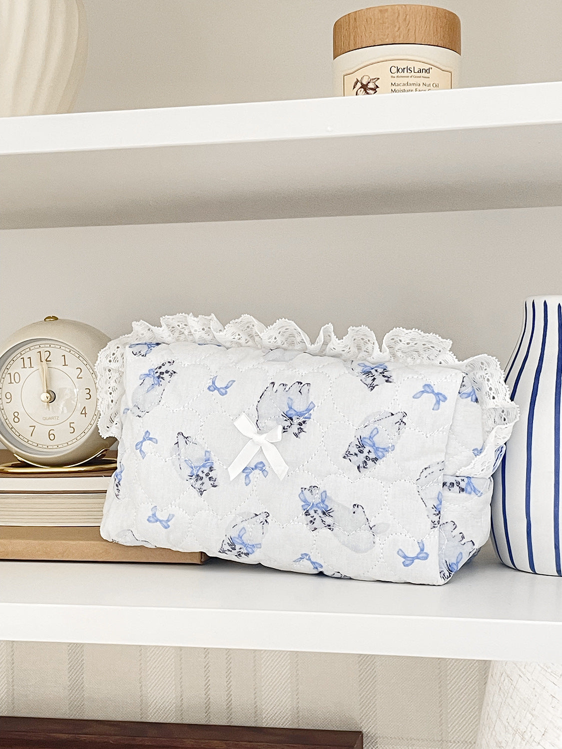 A white cosmetic storage pouch with lace trim and blue floral embroidery rests on a white shelf next to a clock.