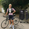 A woman wearing a pink shirt and black cycling shorts stands next to a black hydration vest backpack and a bicycle.