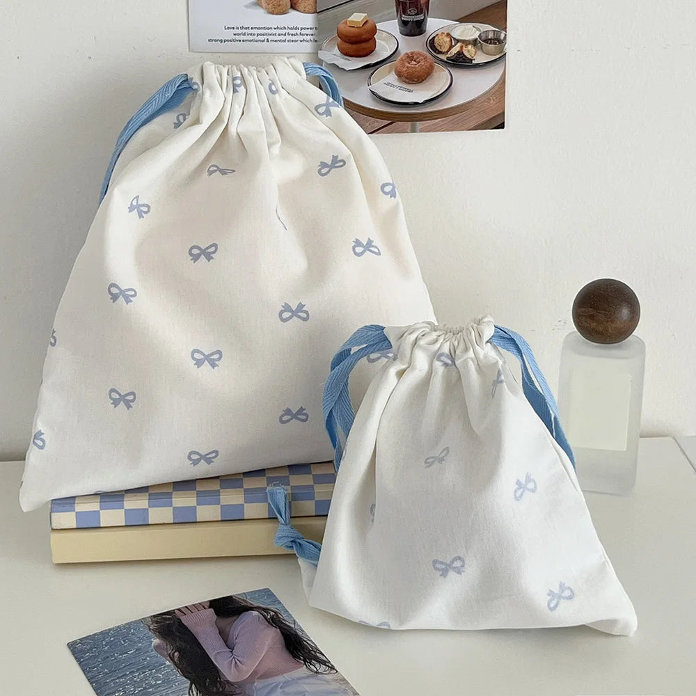 A white canvas makeup bag with light blue bow accents is shown next to a stack of books and a glass bottle.