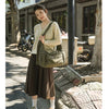 A woman wearing a beige knit sweater and dark skirt holds a large olive green cotton canvas shoulder crossbody bag outdoors.