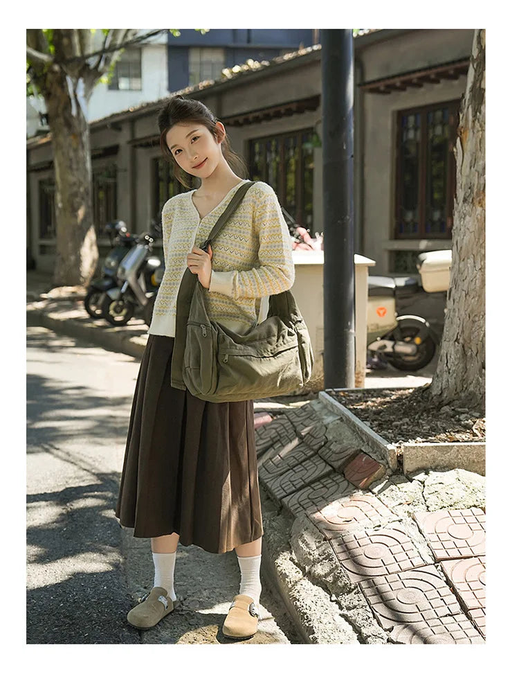 A woman wearing a beige knit sweater and dark skirt holds a large olive green cotton canvas shoulder crossbody bag outdoors.