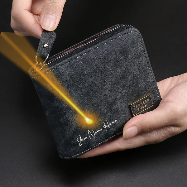 A dark gray leather zipper wallet with personalized engraving on the front is held by a hand, showing a bright light beam.