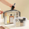 A hand is placing a black bottle into a clear PVC toiletry bag with a zipper closure.