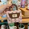 Bolso cruz de textura floral rosa claro con solapa de cuero marrón y correa de cadena dorada, sostenido en la mano.