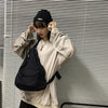 A person wearing a beige sweatshirt and black hat holds a black canvas crossbody bag against a slatted wall.