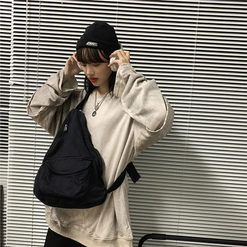 A person wearing a beige sweatshirt and black hat holds a black canvas crossbody bag against a slatted wall.