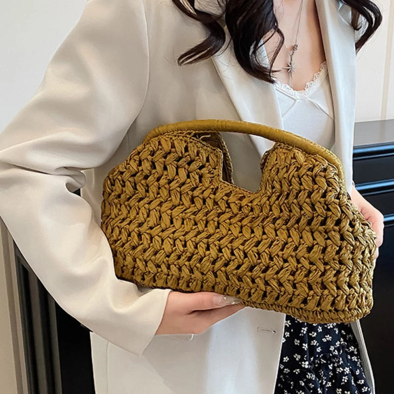 A woman holds a woven straw tote handbag with a light brown color against a white blazer.