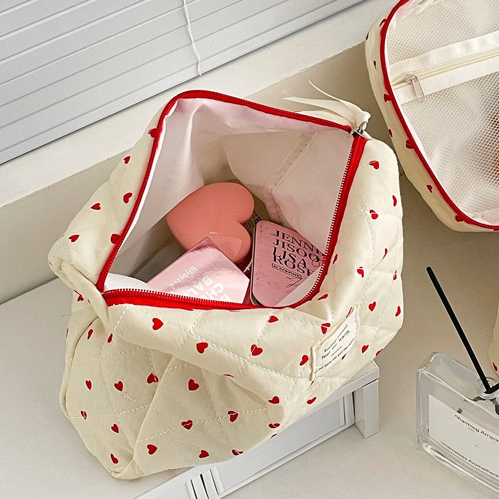 A cream quilted makeup bag with a red zipper and pink lining, featuring a pattern of small red hearts.