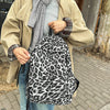 A woman wearing a beige coat and gray sweater carries a leopard print canvas backpack.