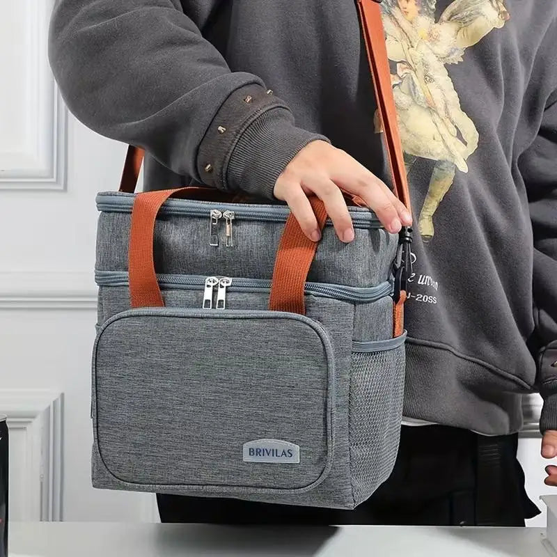 A person holds a grey insulated double-layer lunch bag with brown straps against a white background.