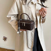A person wearing a beige trench coat holds a brown quilted leather crossbody bag with an adjustable silver chain strap.