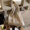 A cream-colored knitted shoulder bag is displayed on a wooden surface next to a glass and other items.