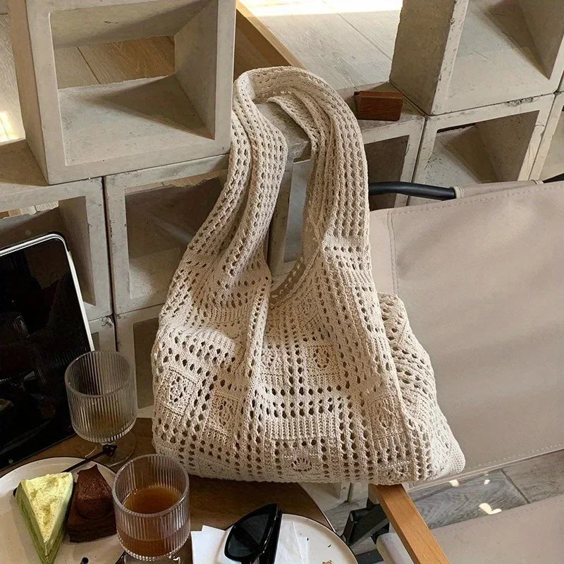 A cream-colored knitted shoulder bag is displayed on a wooden surface next to a glass and other items.