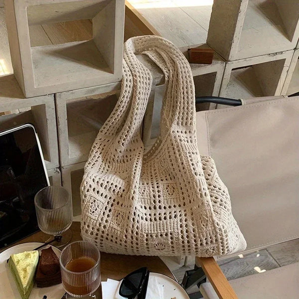 A cream-colored knitted shoulder bag is displayed on a wooden surface next to a glass and other items.