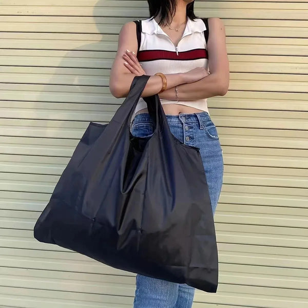 A woman wearing a white top and jeans holds a large black folding tote shopping bag.