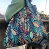 A reusable tote bag featuring a dark floral pattern with teal, pink, and olive green leaves is shown against a wooden surface