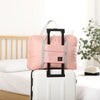 Un sac de voyage pliable rose avec une poignée noire est posé sur une valise à roulettes blanche.