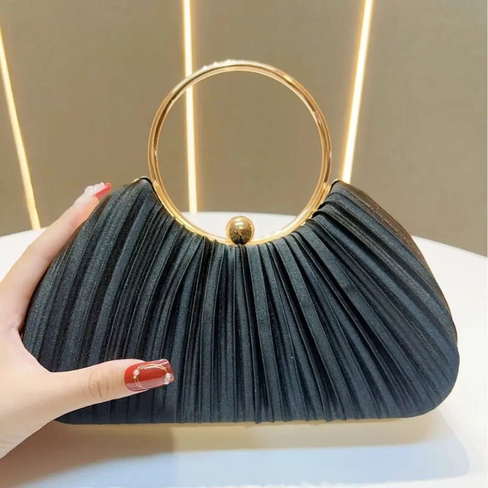 A black satin pleated evening clutch bag with a gold hoop and stud detail is held by a hand.
