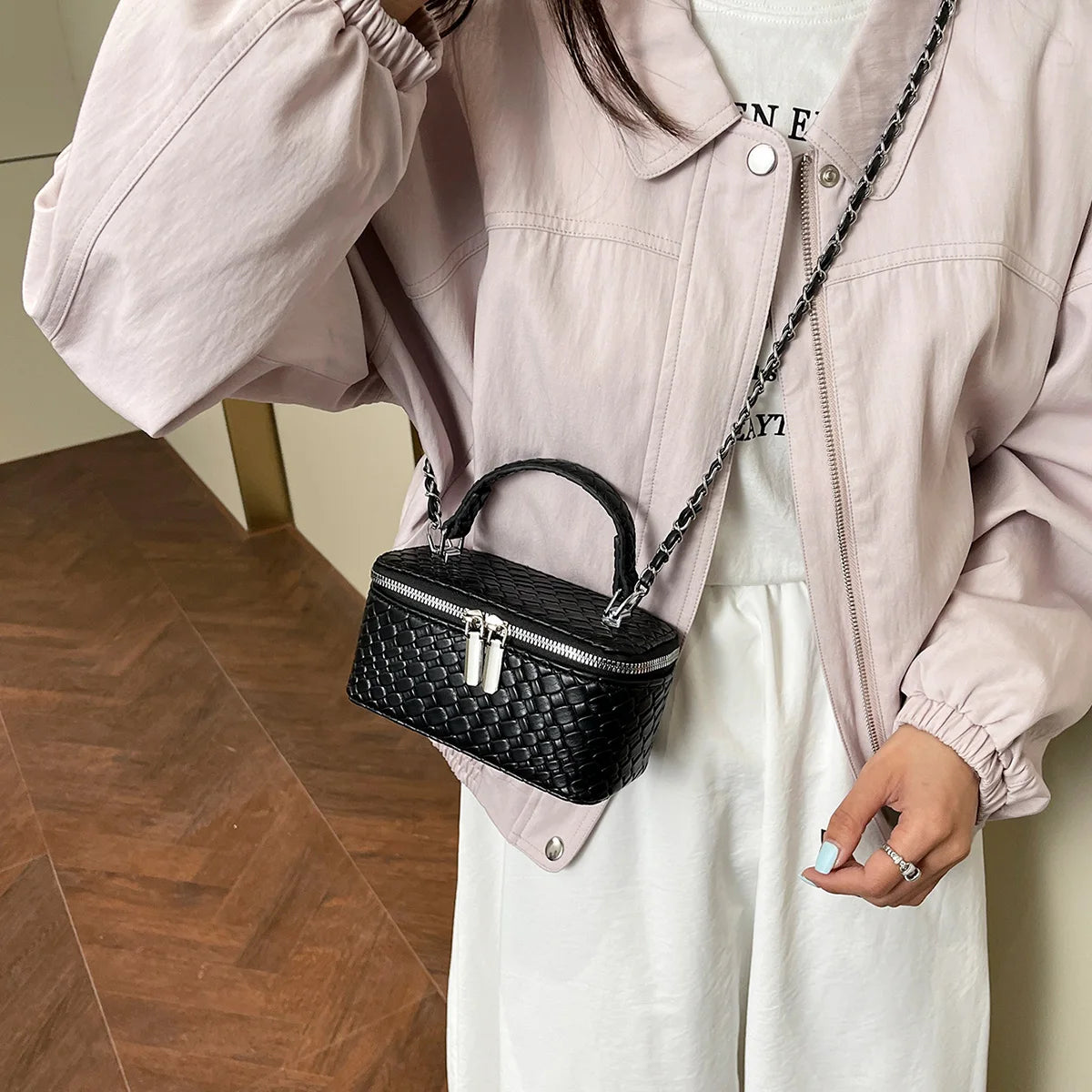 A person wearing a light pink jacket and white top holding a black woven crossbody bag with a silver chain strap.