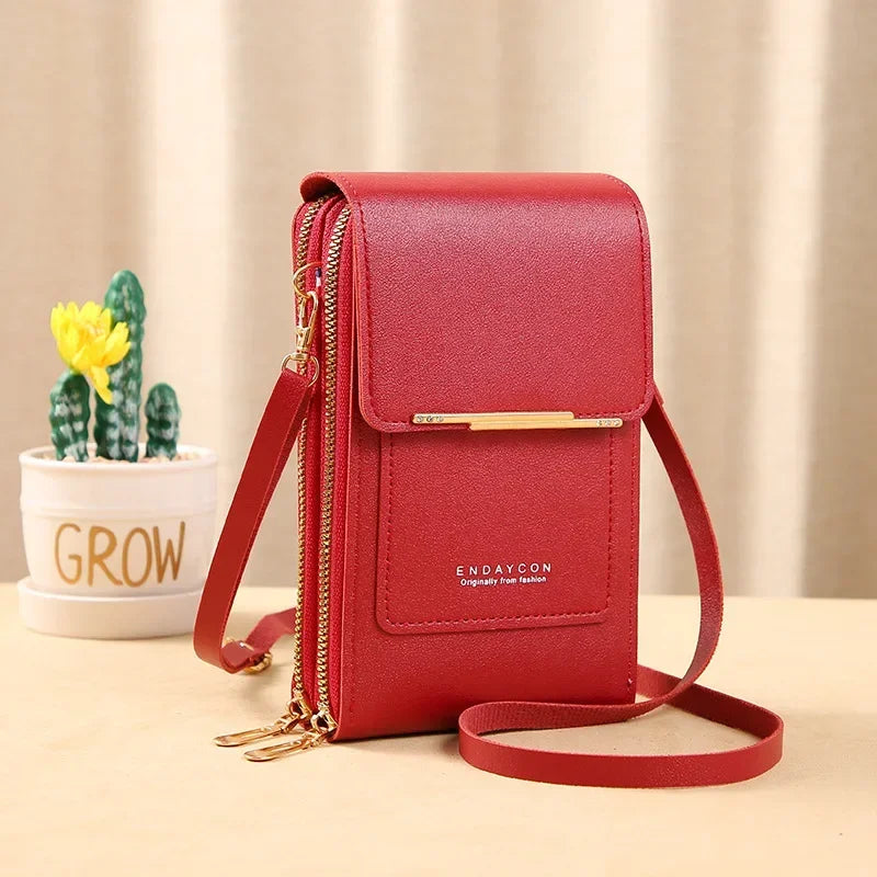 A red leather women's touch screen crossbody shoulder bag with gold hardware resting on a light wooden surface.