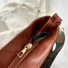 A brown leather shoulder bag with a gold zipper and an embroidered bohemian strap is shown against a white fabric background.