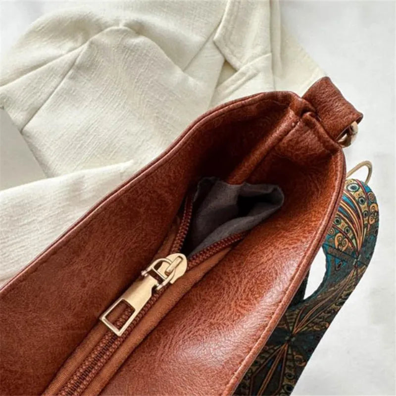 A brown leather shoulder bag with a gold zipper and an embroidered bohemian strap is shown against a white fabric background.