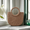 A natural beige handwoven rattan beach bag resting on a white windowsill next to a window frame.
