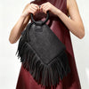 A woman holds a black fringed clutch bag with tassel detail against a dark red dress.