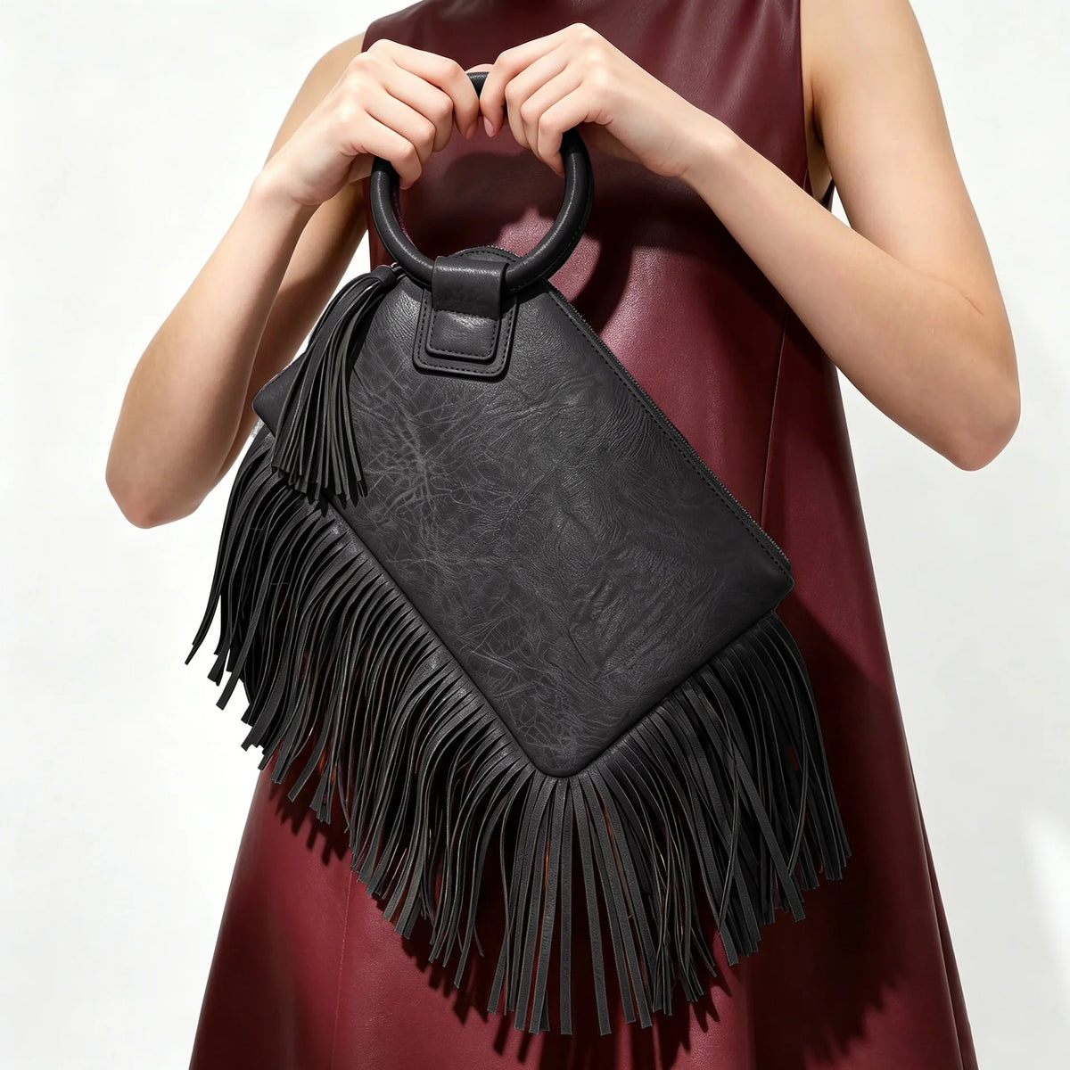 A woman holds a black fringed clutch bag with tassel detail against a dark red dress.