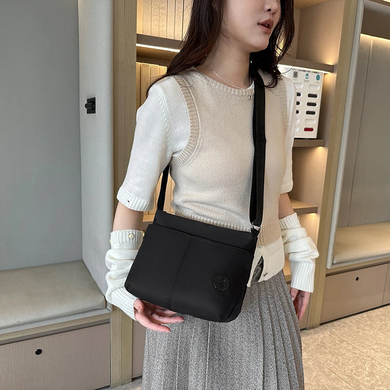 A woman wearing a cream knit top and gray skirt holds a black waterproof nylon crossbody bag.
