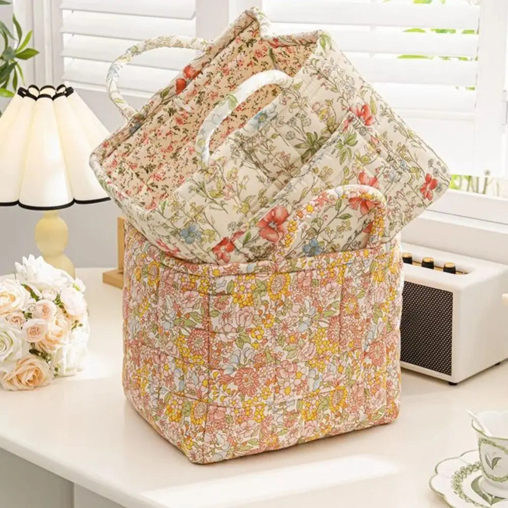 A quilted fabric storage basket with carry handles featuring a floral pattern, resting on a white surface near a window.
