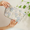 A hand holds a light blue floral canvas makeup bag with gold embroidery against a white background.