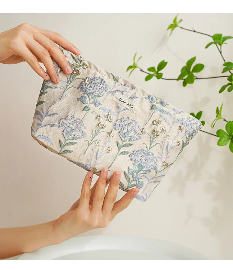 A hand holds a light blue floral canvas makeup bag with gold embroidery against a white background.