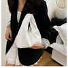 A woman holds a white minimalist crossbody tote bag against a black blazer and a white skirt.