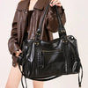 A woman holds a black vintage washed leather tote bag with gold hardware and a brown leather jacket.