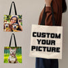 A person holds a beige canvas tote bag with black handles, featuring text that reads Custom Your Picture.