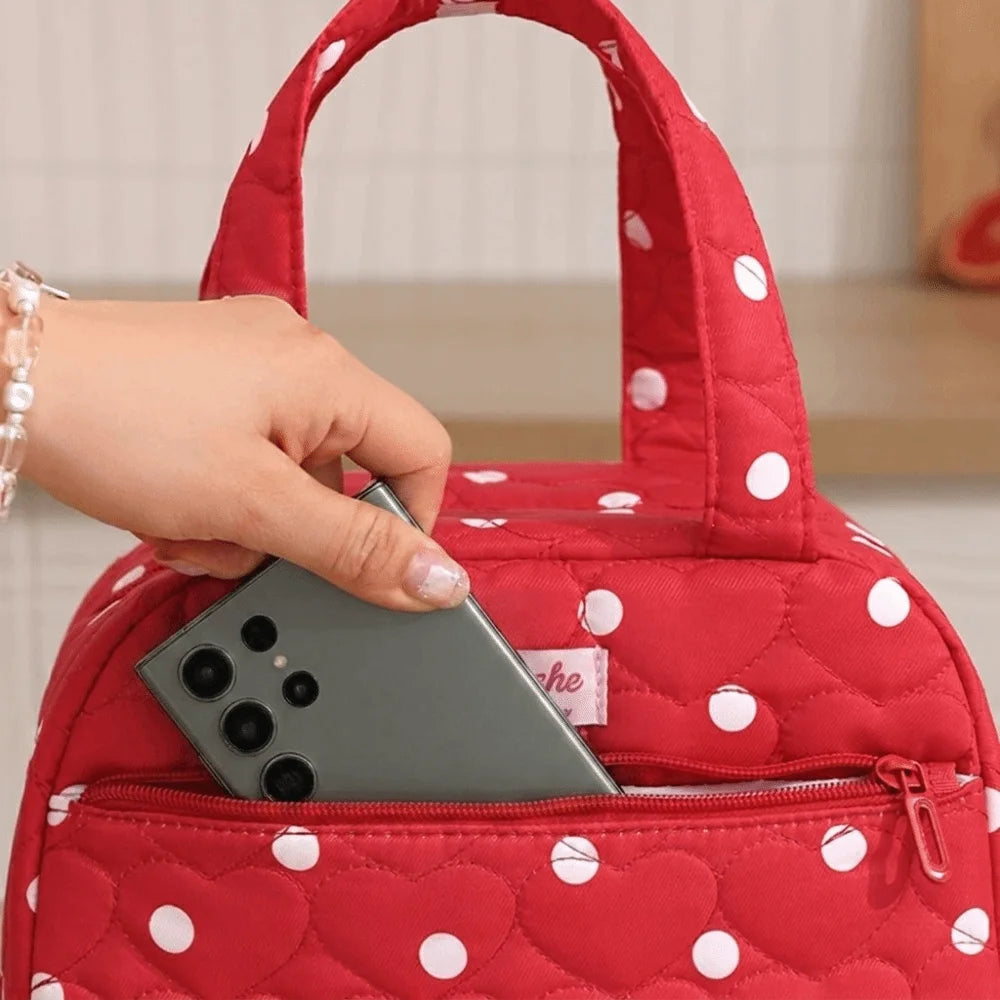 A hand places a smartphone into the pocket of a red quilted lunch tote bag with white polka dots.