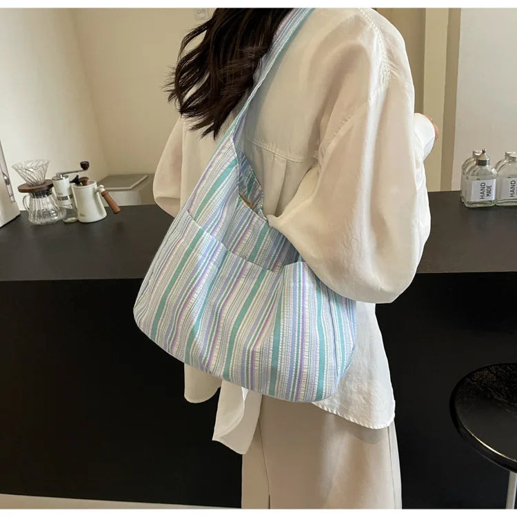 A light striped canvas shoulder bag with a magnetic closure is displayed next to a white garment.
