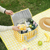 A hand is opening a yellow insulated picnic tote filled with food items on a yellow checkered blanket.