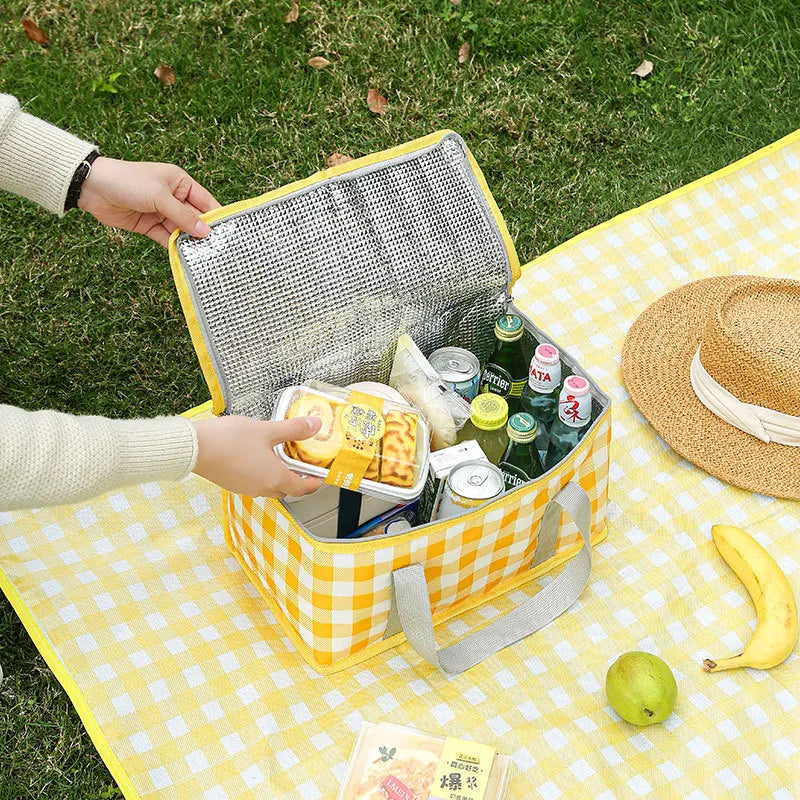 A hand is opening a yellow insulated picnic tote filled with food items on a yellow checkered blanket.