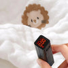A hand holds a black electronic device with a red display showing 10:00 and a light brown lion face printed on white fabric.