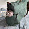 A sage green nylon tote bag with a brown strap, featuring an interior pocket holding rolled pink fabric.