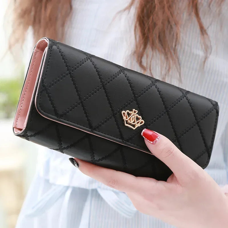 A black quilted long wallet with a gold crown buckle is held by a hand against a light background.